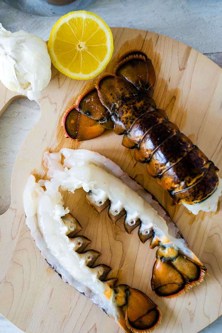 Whole lobster tail and split lobster tail on cutting board with garlic and lemon