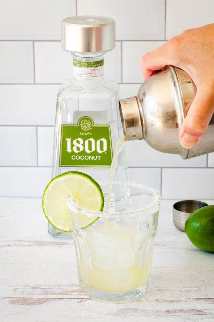 Cocktail shaker pouring margarita into glass with lime and salt on rim, 1800 coconut tequila