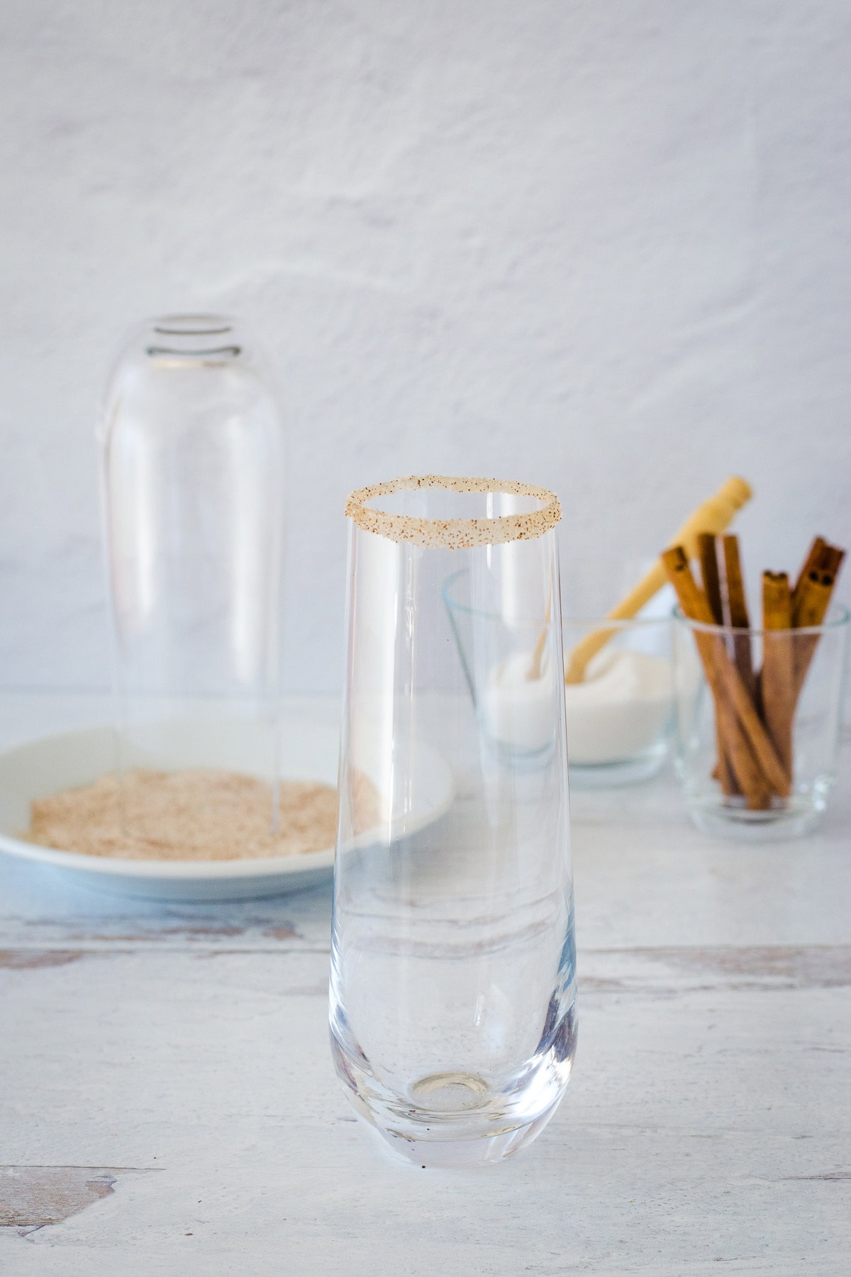 Stemless champagne flute rimmed with cinnamon sugar with another flute upside down on plate and sugar and cinnamon sticks in background. 