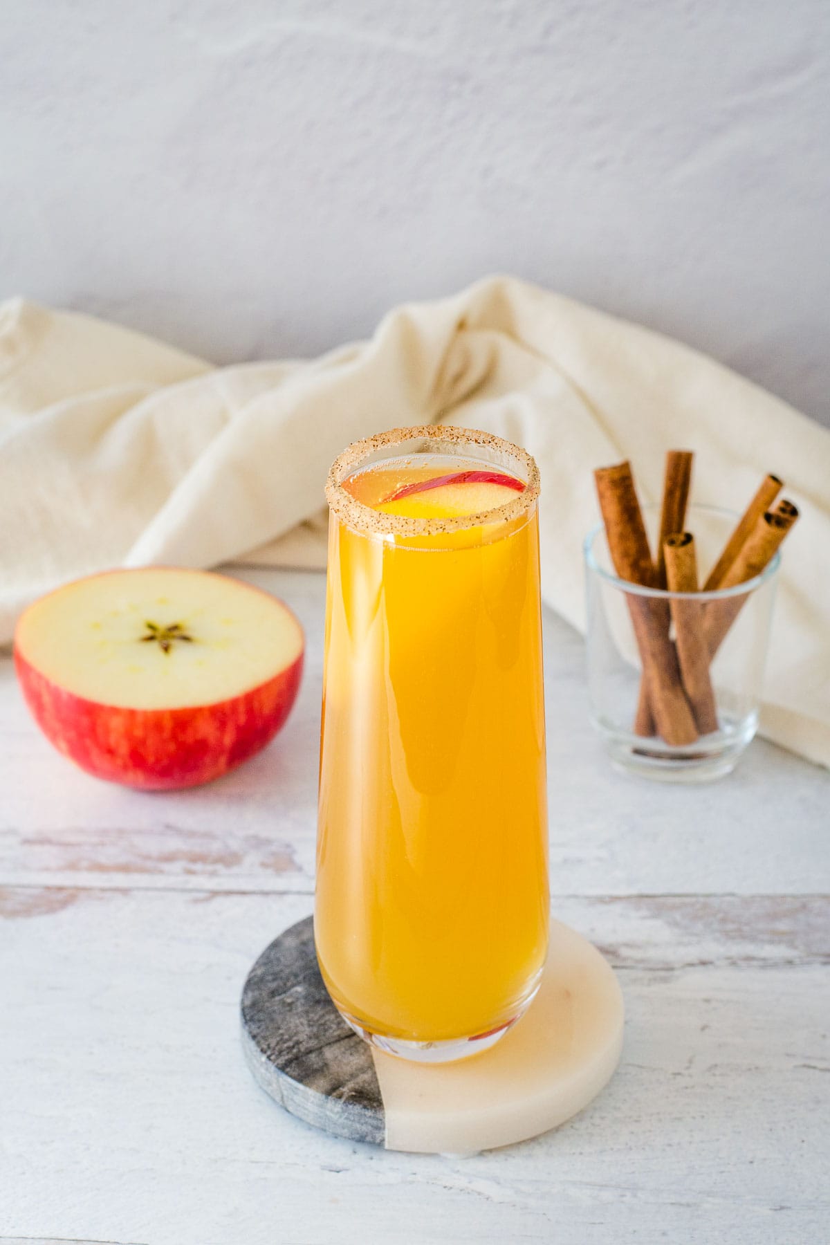 Apple cider mimosa in stemless champagne flute rimmed with cinnamon sugar and garnished with a red apple slice, with apple and cinnamon sticks in the background.