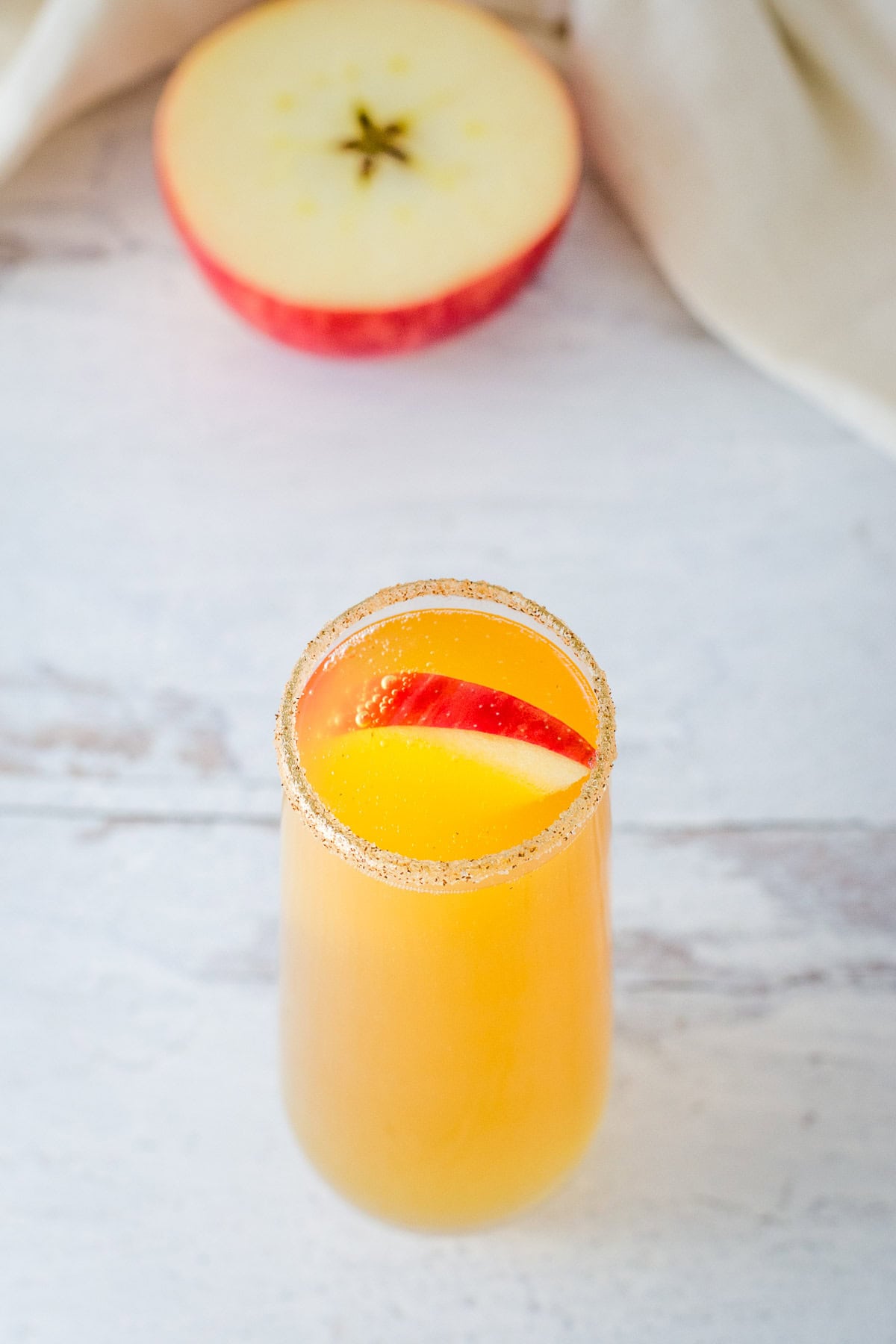 Apple cider mimosa in stemless champagne flute rimmed with cinnamon sugar and garnished with a red apple slice, with apple half and cream napkin in the background.
