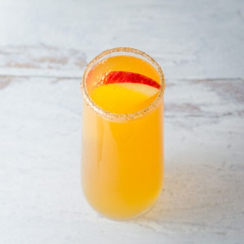 Apple cider mimosa in stemless champagne flute rimmed with cinnamon sugar and garnished with a red apple slice.