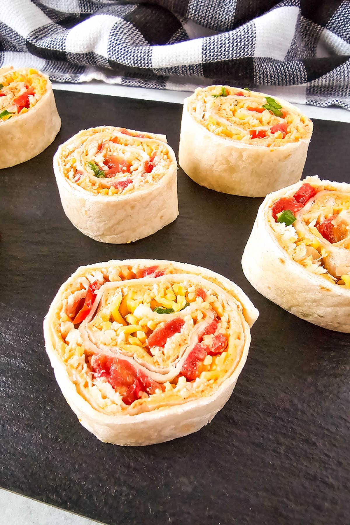 Five chicken pinwheels filled with colorful ingredients like chopped vegetables and cheese are displayed on a dark serving board.