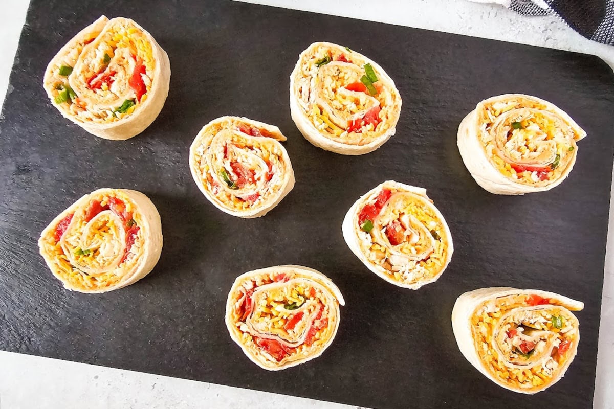 Eight chicken taco pinwheels filled with colorful ingredients like chopped vegetables and cheese are displayed on a dark serving board.