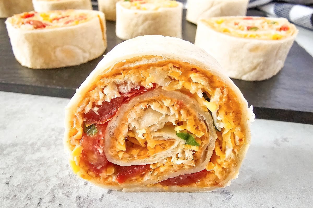 Taco pinwheel filled with chicken, vegetables and cheese is standing on a white counter with more pinwheels in the background on a black board.