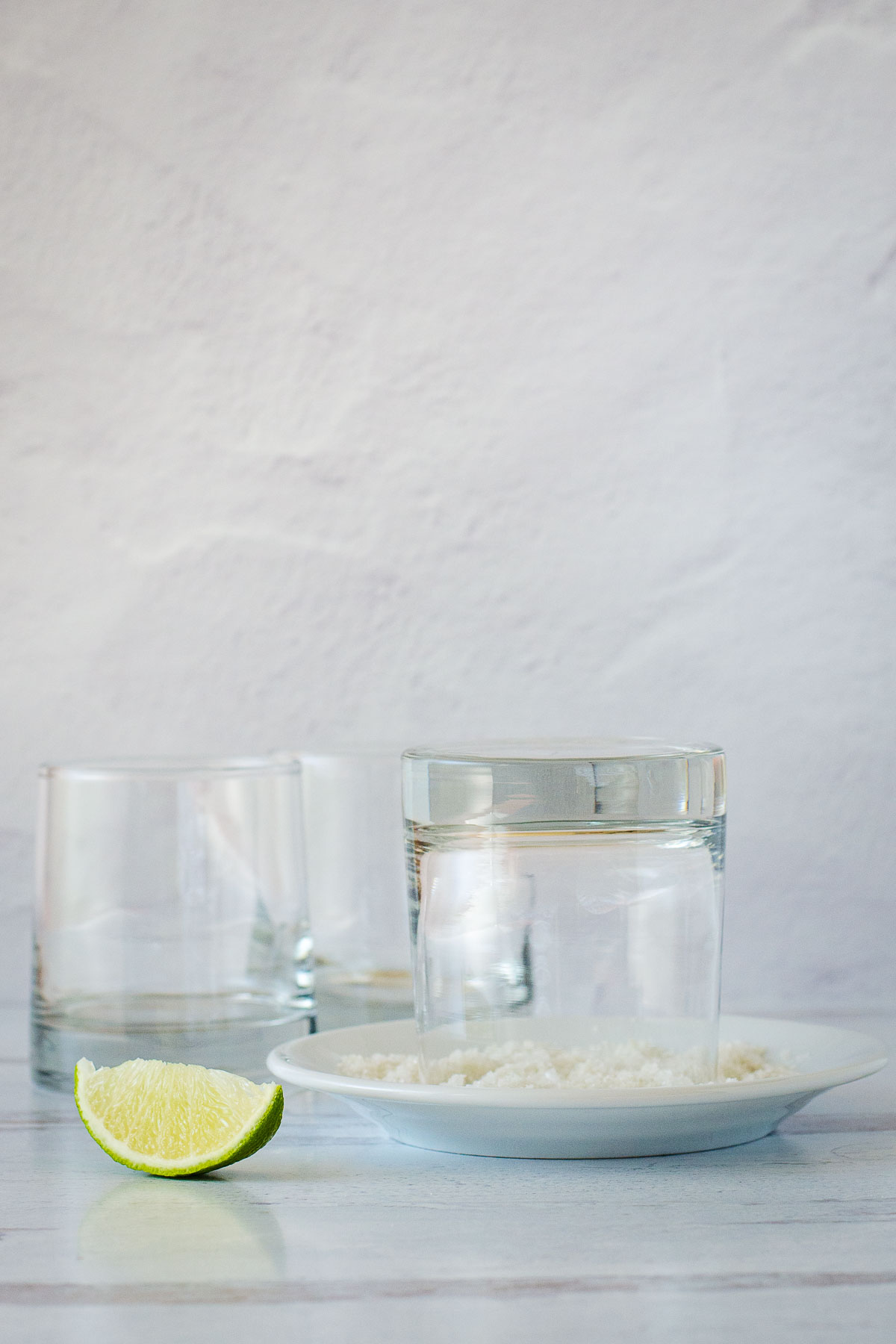 Rocks glass upside down in plate of coarse salt with lime wedge in foreground and two glasses in background.