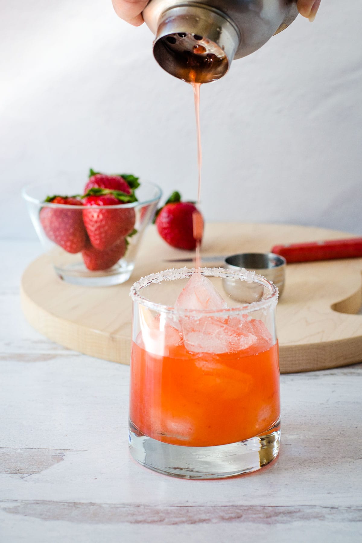 Pouring pink drink from shaker into glass with strawberries and paring knife on cutting board in background.