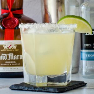 Classic margarita with lime wedge on rim of glass, bottles and shaker in background.