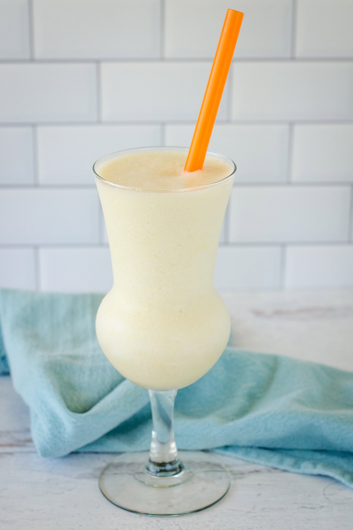 Frozen pina colada in tall glass with orange straw and blue towel in background.