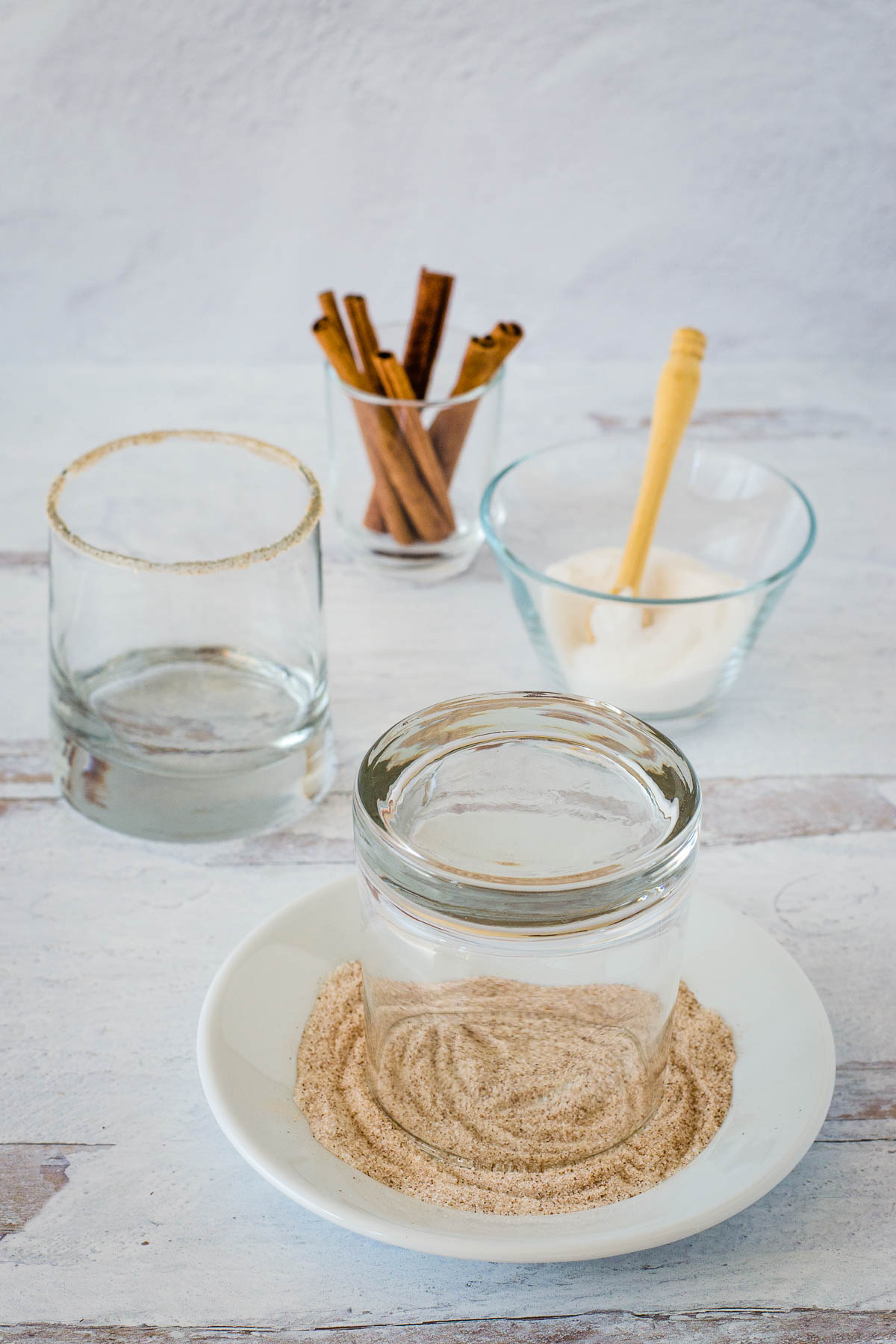 Rocks glass upside down in small plate of spice and sugar mixture with rimmed glass, cinnamon sticks and a dish of sugar in the background.