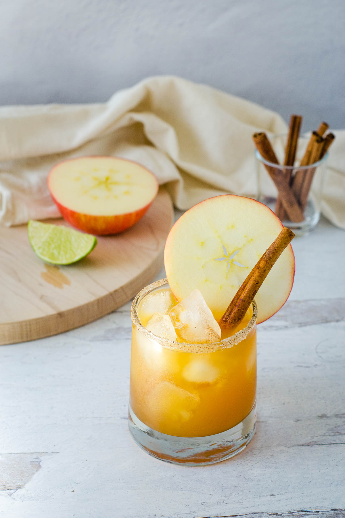 Apple cider margarita with sugar spice rim, apple slice and cinnamon stick with apple half, lime wedge and cinnamon sticks in background.