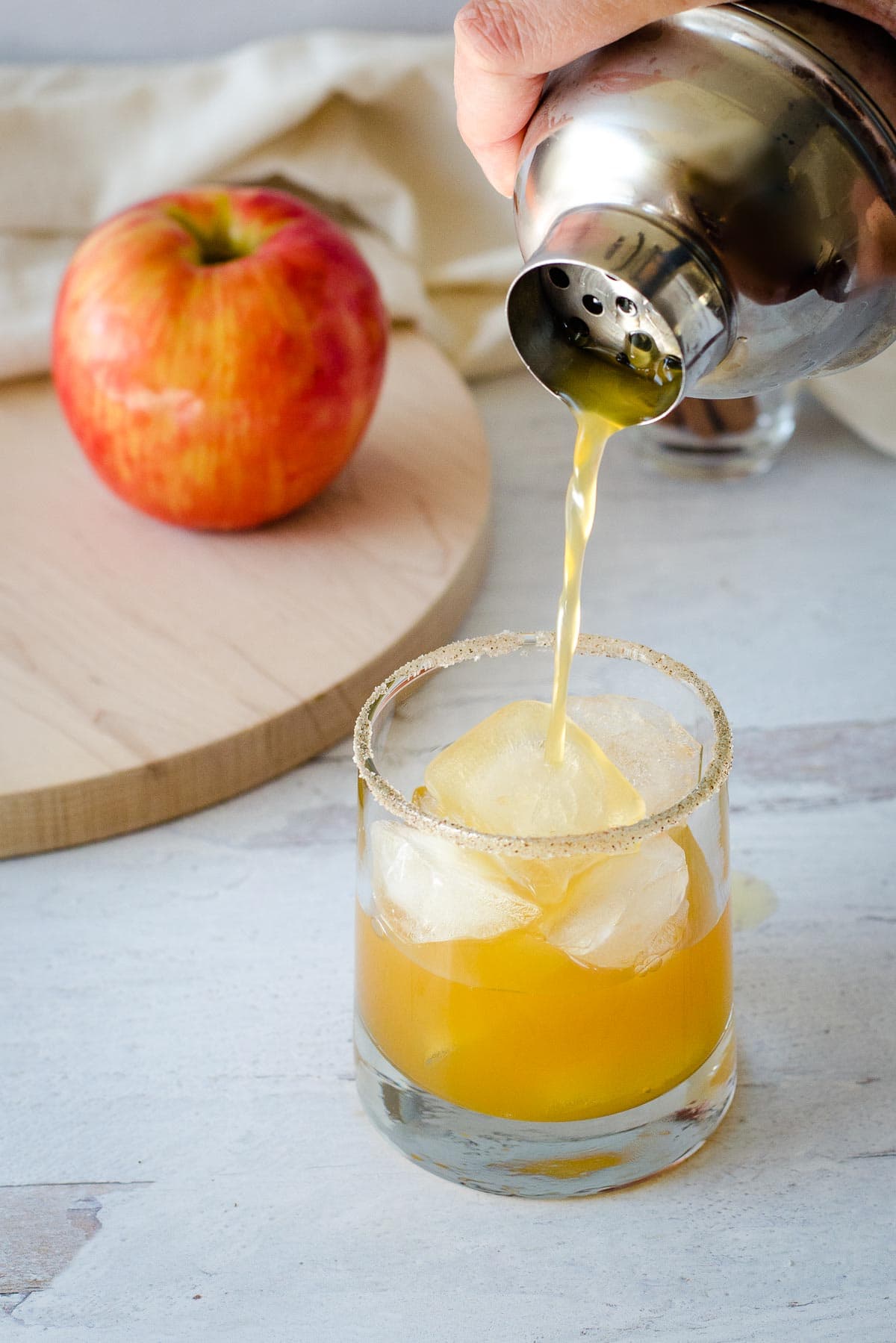 Cocktail shaker pouring drink into rimmed glass with ice cubes, apple on cutting board in background.