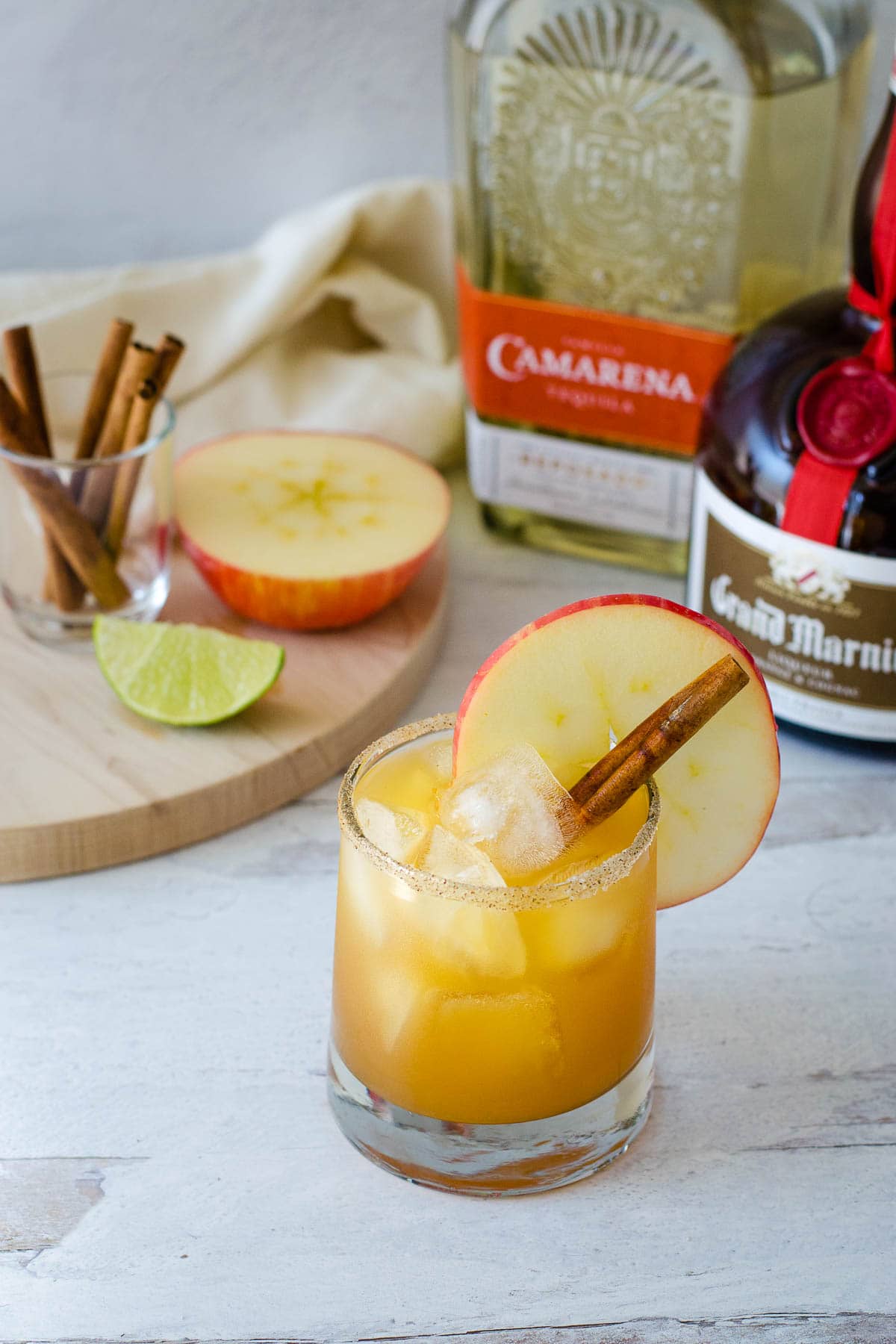 Apple cider margarita with sugar spice rim, apple slice and cinnamon stick with apple half, lime wedge and cinnamon sticks, tequila and Grand Marnier in background.