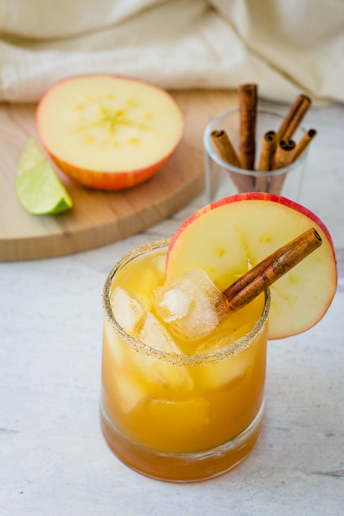 Apple cider margarita with sugar spice rim, apple slice and cinnamon stick with apple half, lime wedge and cinnamon sticks in background.