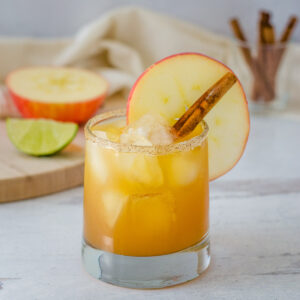 Apple cider margarita with sugar spice rim, apple slice and cinnamon stick with apple half, lime wedge and cinnamon sticks in background.