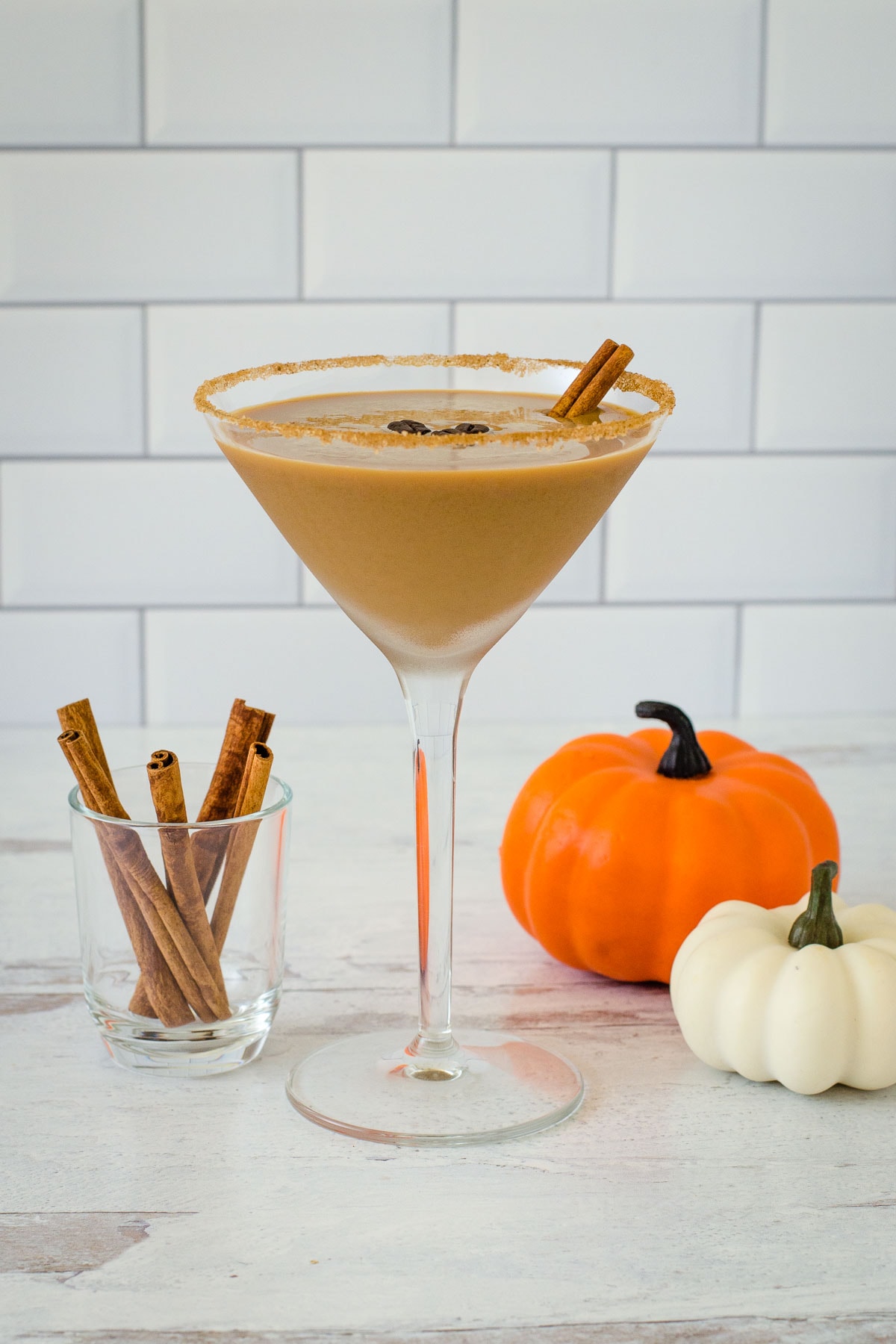 Pumpkin spice espresso martini with three coffee beans and cinnamon stick garnish, cinnamon sticks and pumpkins in background.