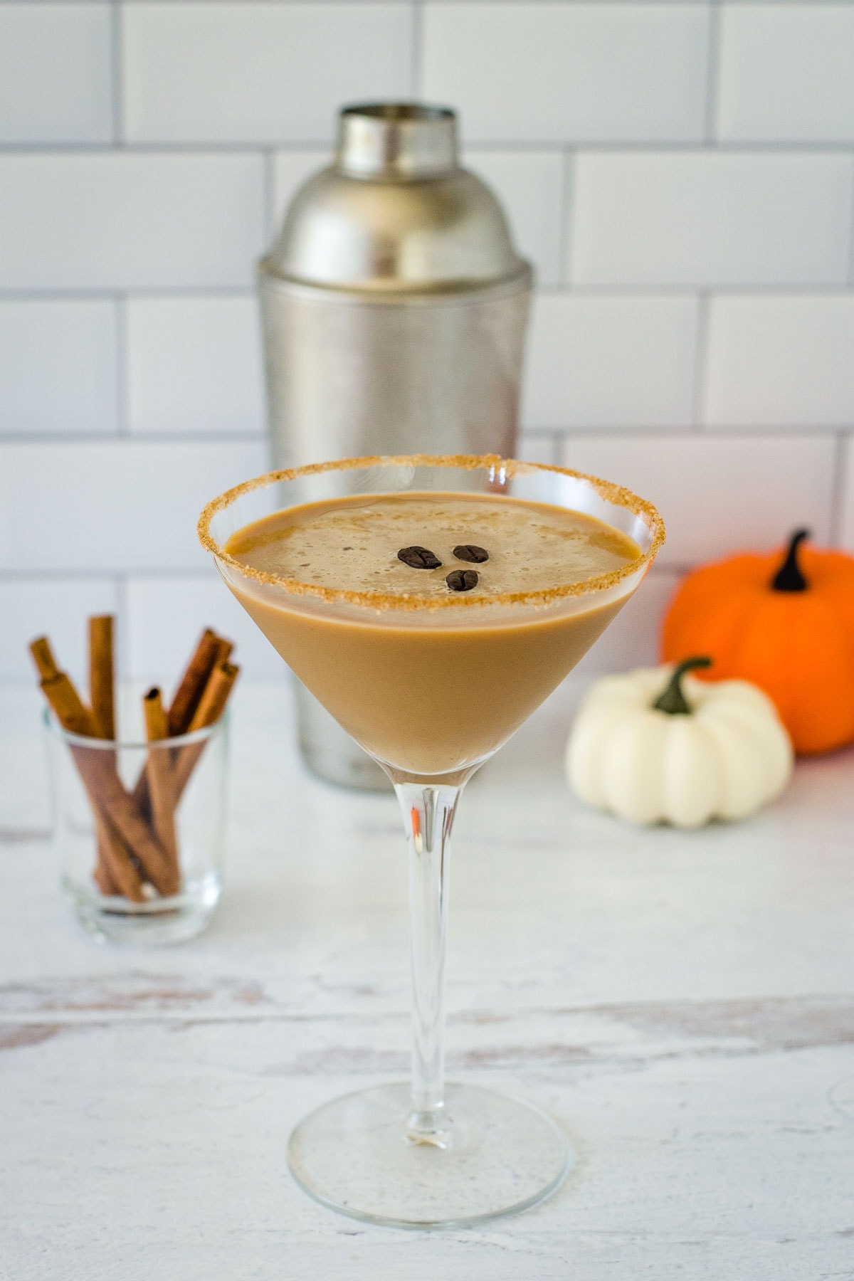Pumpkin spice espresso martini with three coffee beans and cinnamon stick garnish, cinnamon sticks and pumpkins in background.