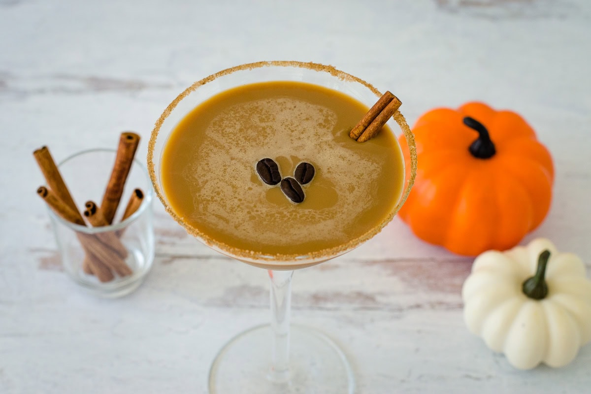 Pumpkin spice espresso martini with three coffee beans and cinnamon stick garnish, cinnamon sticks, orange and white min pumpkins in background.