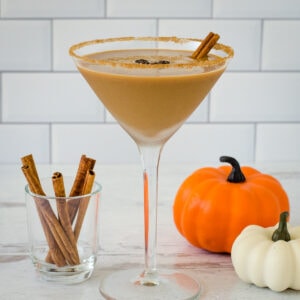 Pumpkin spice espresso martini with three coffee beans and cinnamon stick garnish, cinnamon sticks and pumpkins in background.