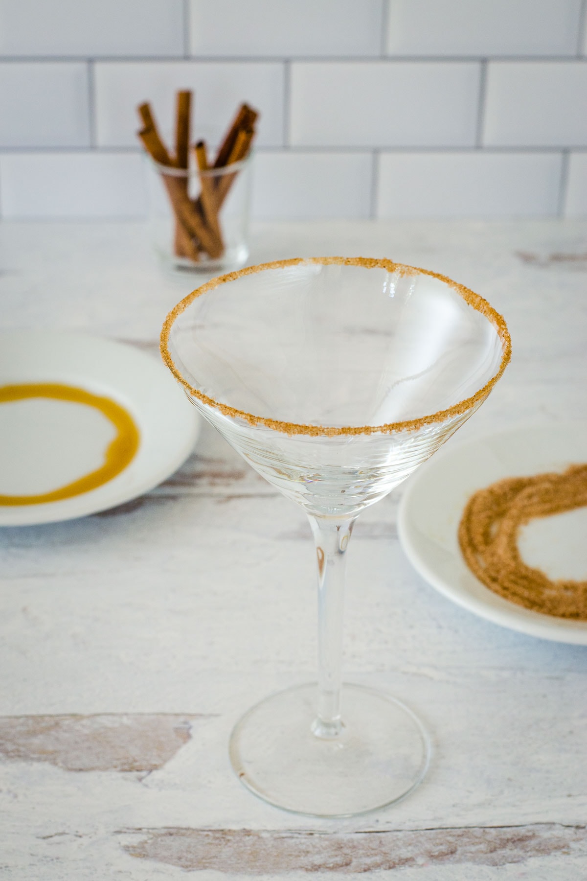 Cocktail glass rimmed with spiced sugar, cinnamon sticks and small plates with maple syrup and spice mixture in backgrouns.