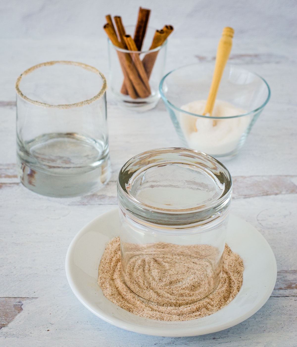 Short glass upside down in a dish of pumpkin spice mixed with sugar with a sugar spice rimmed glass, cinnamon sticks and a bowl of sugar in the background.