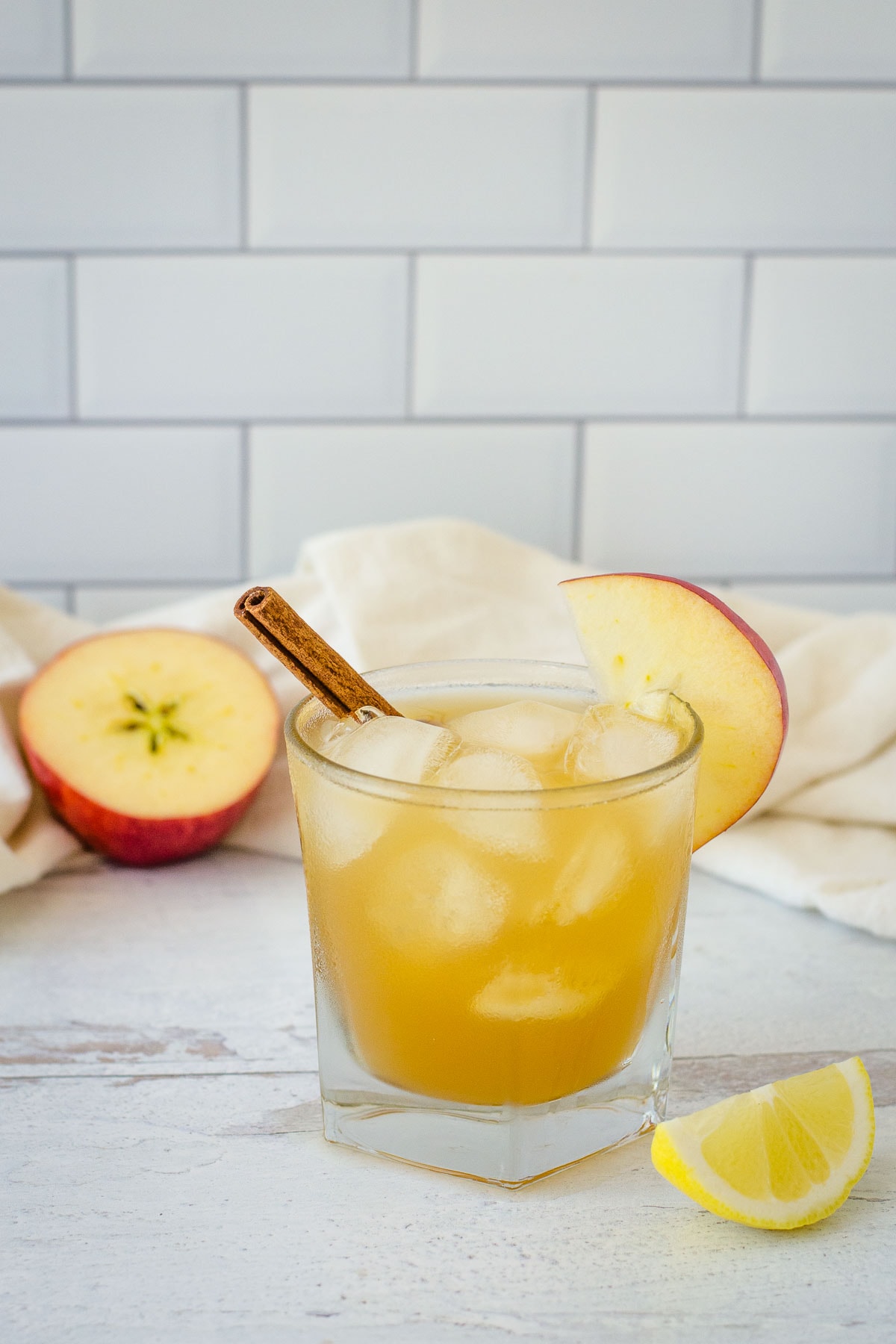 Apple cider bourbon cocktail garnished with apple slice and cinnamon stick next to apple half, lemon wedge and cream towel.