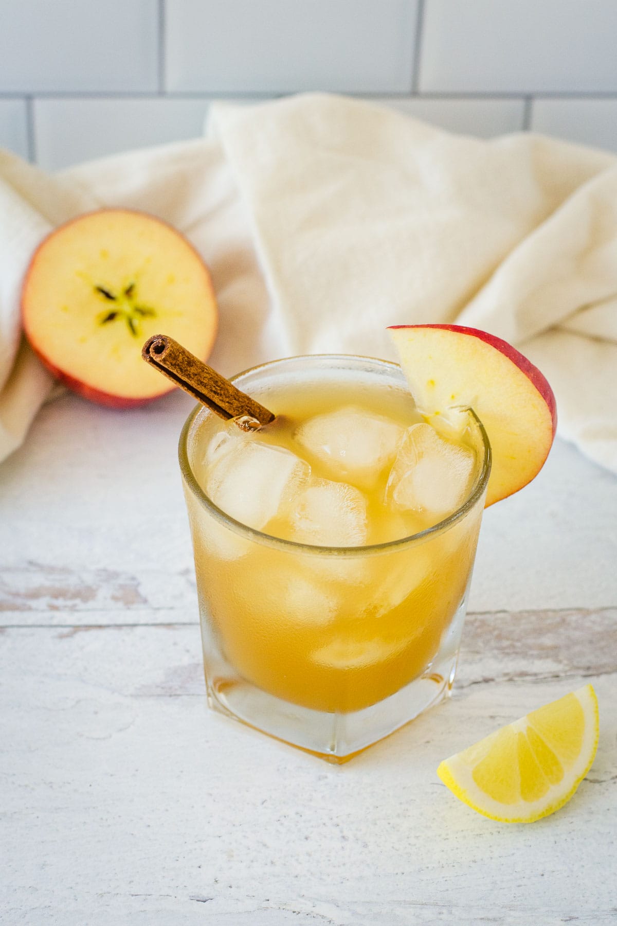 Apple cider bourbon cocktail garnished with apple slice and cinnamon stick next to apple half, lemon wedge and cream towel.