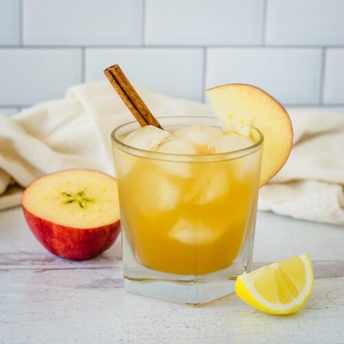 Apple cider bourbon cocktail garnished with apple slice and cinnamon stick next to apple half, lemon wedge and cream towel.