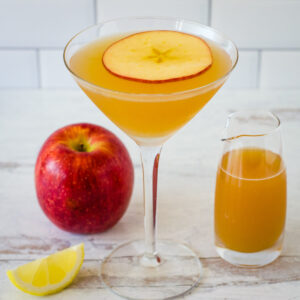 Apple cider martini garnished with apple slice on top and lemon wedge, apple and small pitcher of cider in background.