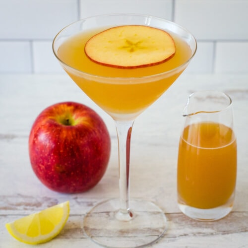 Apple cider martini garnished with apple slice on top and lemon wedge, apple and small pitcher of cider in background.
