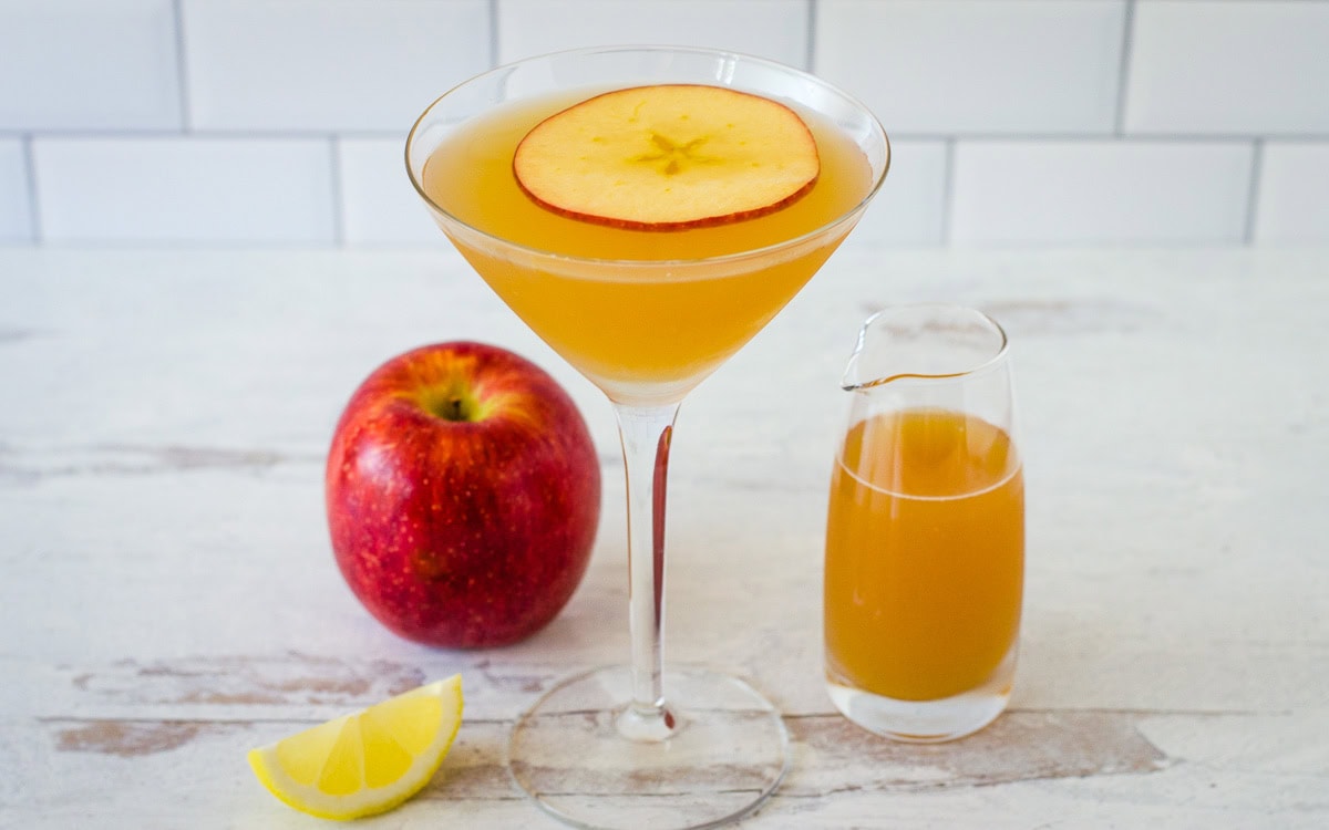 Apple cider martini garnished with apple slice on top and lemon wedge, apple and small pitcher of cider in background.