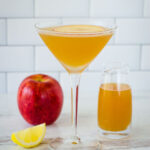 Apple cider martini with apple slice on top next to lemon wedge, apple and small pitcher of cider..