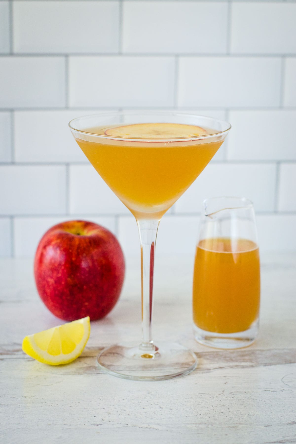 Apple cider martini with apple slice on top next to lemon wedge, apple and small pitcher of cider..
