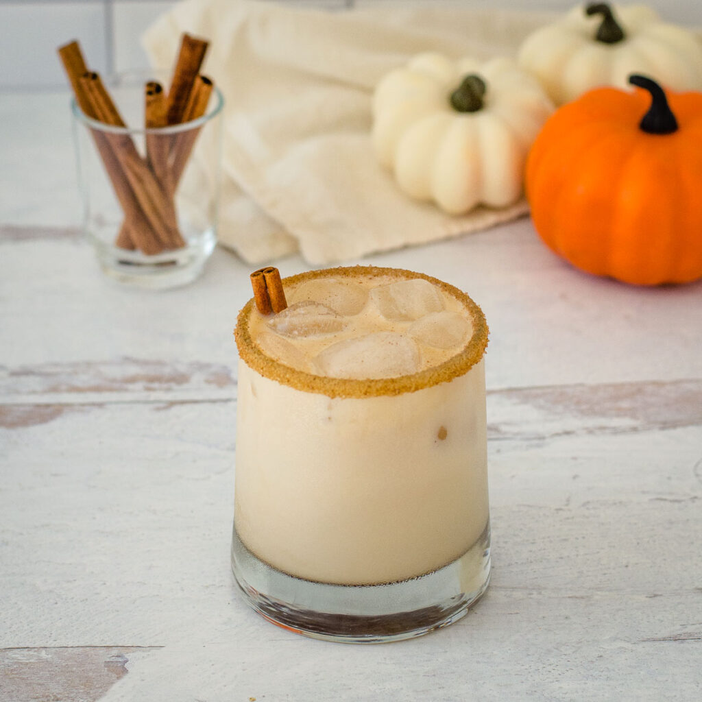 Pumpkin spice white Russian cocktail with cinnamon sticks and mini pumpkins in the background.