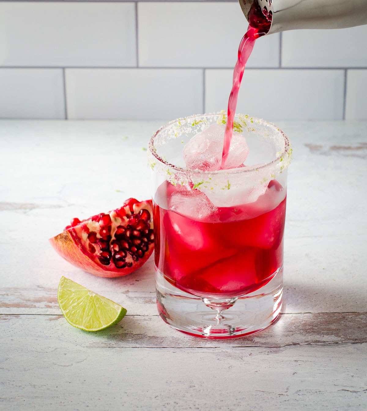 Pouring red drink over ice in glass with pomegranate and lime next to it.