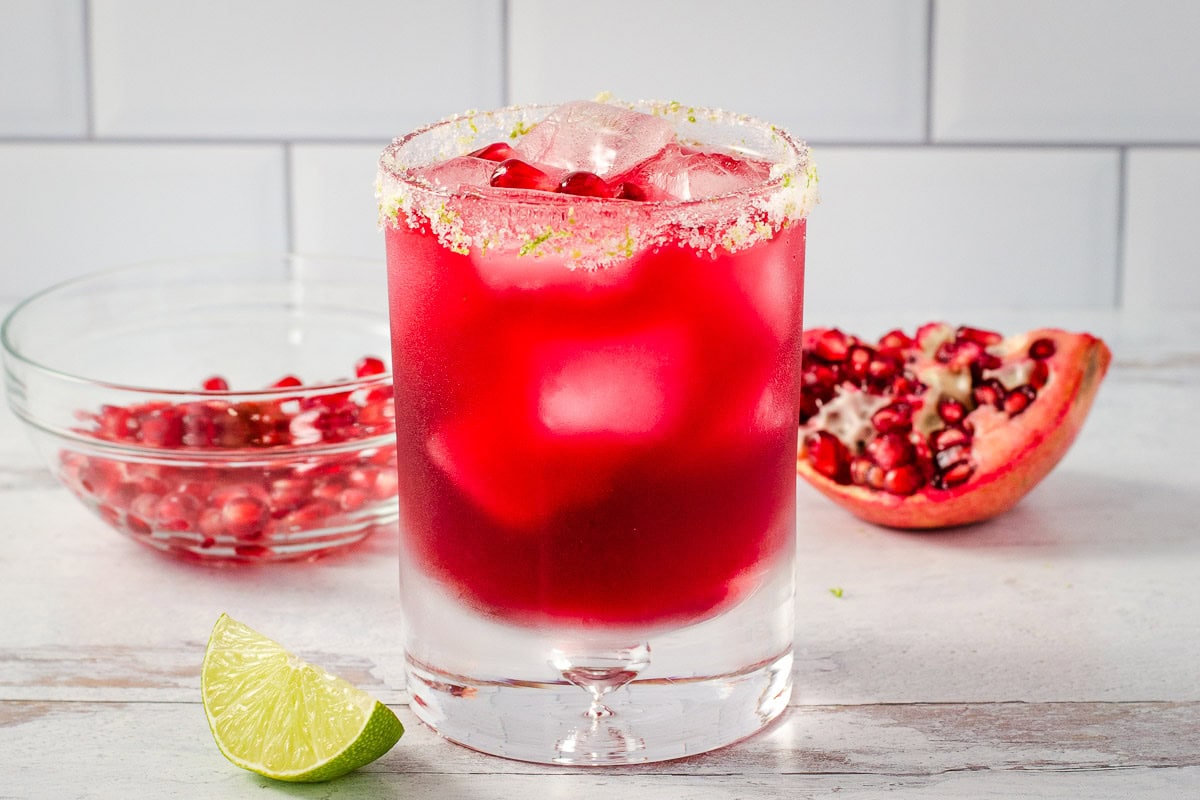 Pomegranate margarita with salted rim and lime next to pomegranate wedge and bowl of seeds.