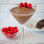 Chocolate raspberry martini with fresh raspberry skewer next to raspberries and small plate with chocolate.