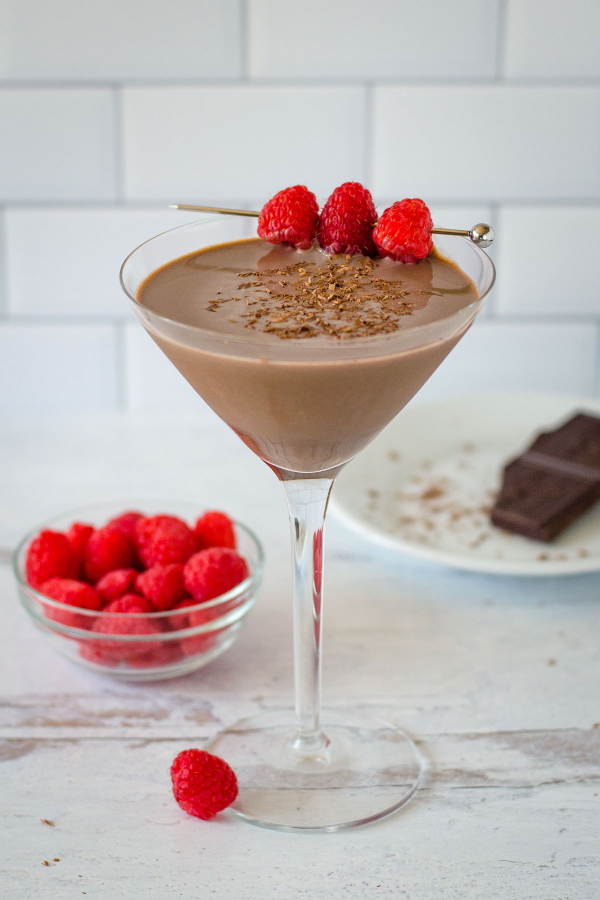 Chocolate raspberry martini with fresh raspberry skewer next to raspberries and small plate with chocolate.