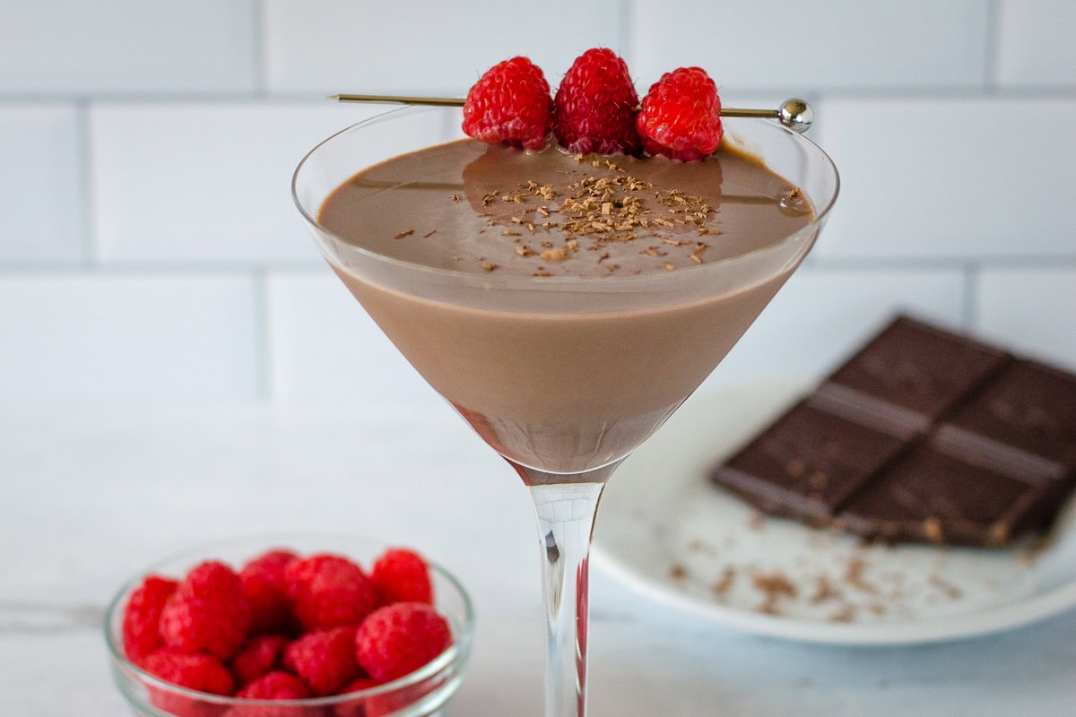Chocolate raspberry martini with fresh raspberry skewer next to bowl of raspberries and small plate of chocolate.