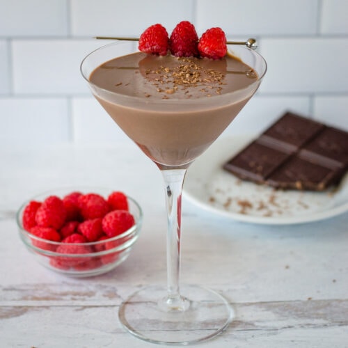 Chocolate raspberry martini with fresh raspberry skewer next to bowl of raspberries and small plate of chocolate.