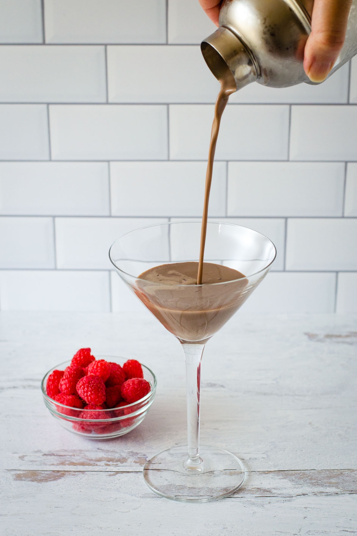 Pouring drink from shaker into cocktail glass with small bowl of raspberries in background.