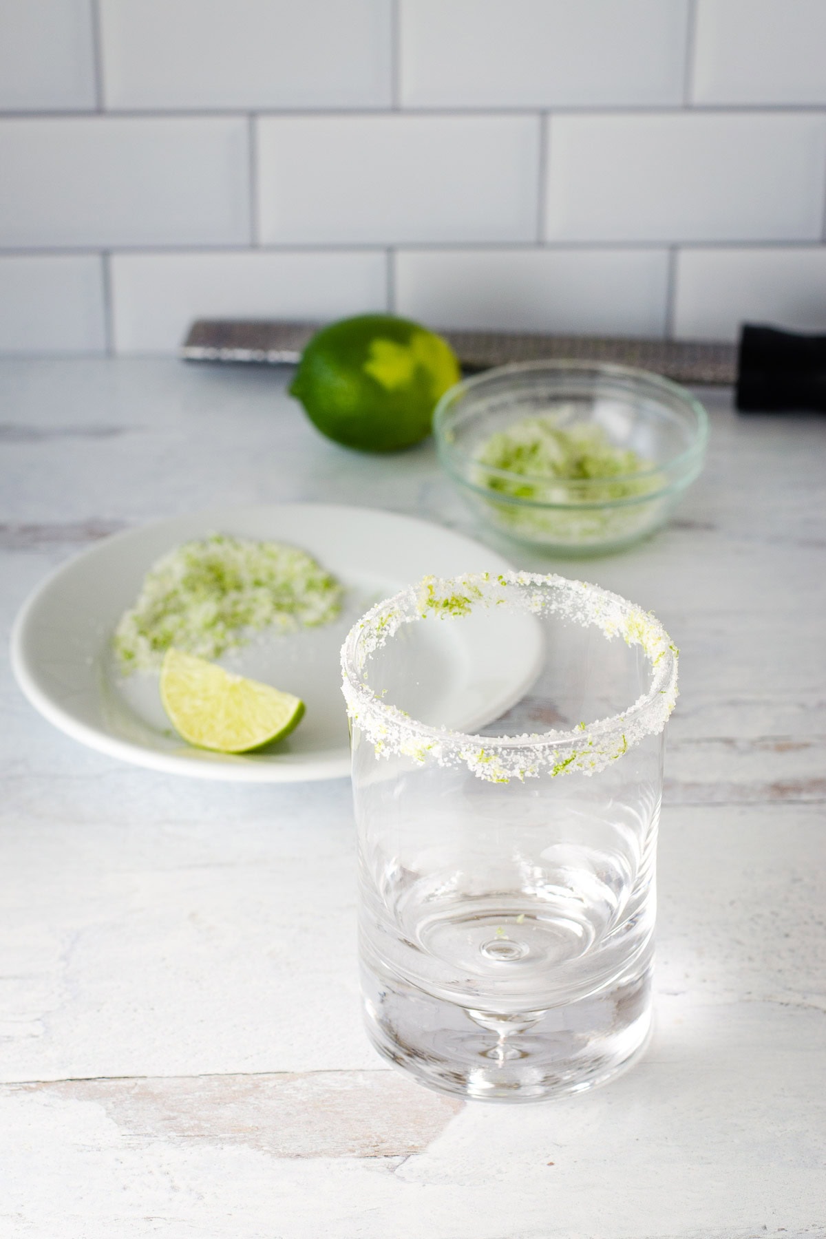 Glass with lime salt rim and limes, zester and dish and bowl of lime salt in background.