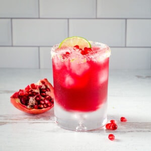 Pomegranate margarita with salted rim and lime next to pomegranate wedge and seeds.