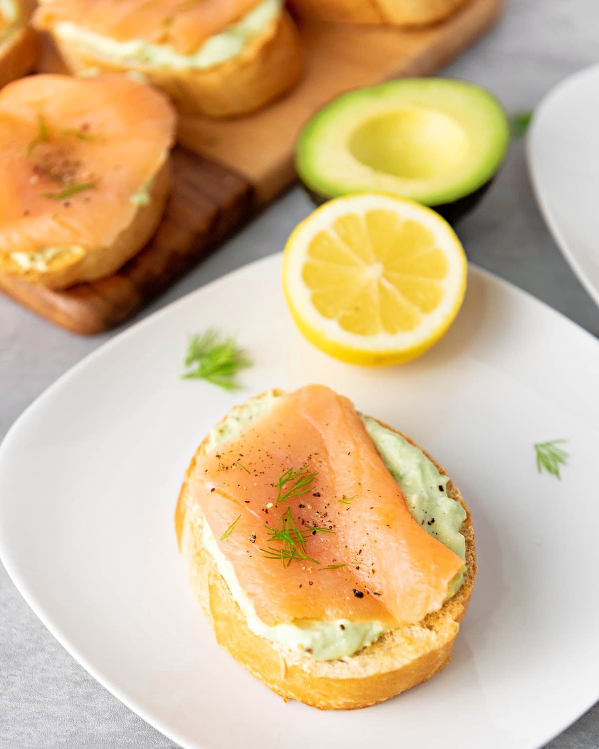 Smoked salmon crostini with avocado cream and fresh dill and board with more appetizers next to avocado and lemon.