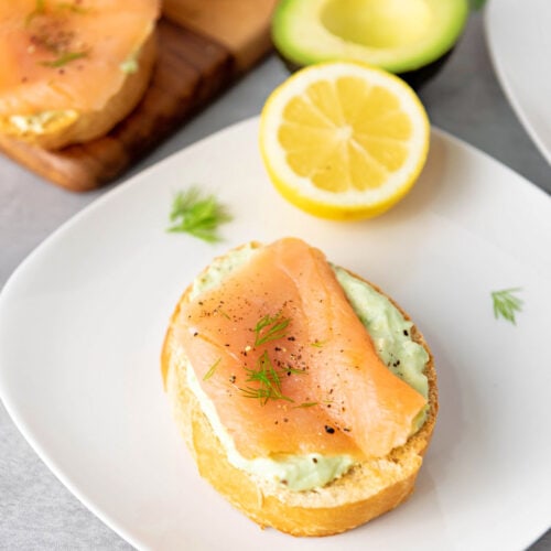 Smoked salmon crostini with avocado cream and fresh dill and board with more appetizers next to avocado and lemon.