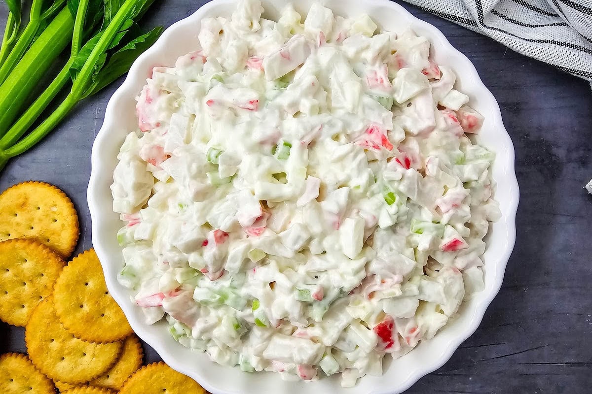 Crab salad in white dish next to crackers, celery stalks and napkin.