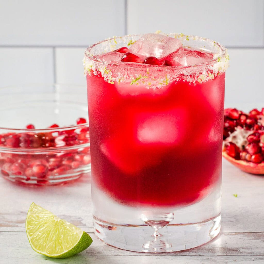 Pomegranate margarita with salted rim and lime next to pomegranate wedge and bowl of seeds.