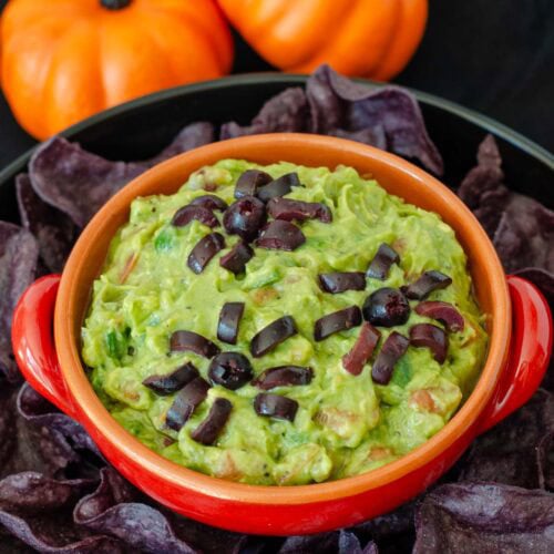 Halloween guacamole with black olive spiders in an orange bowl on a black background.